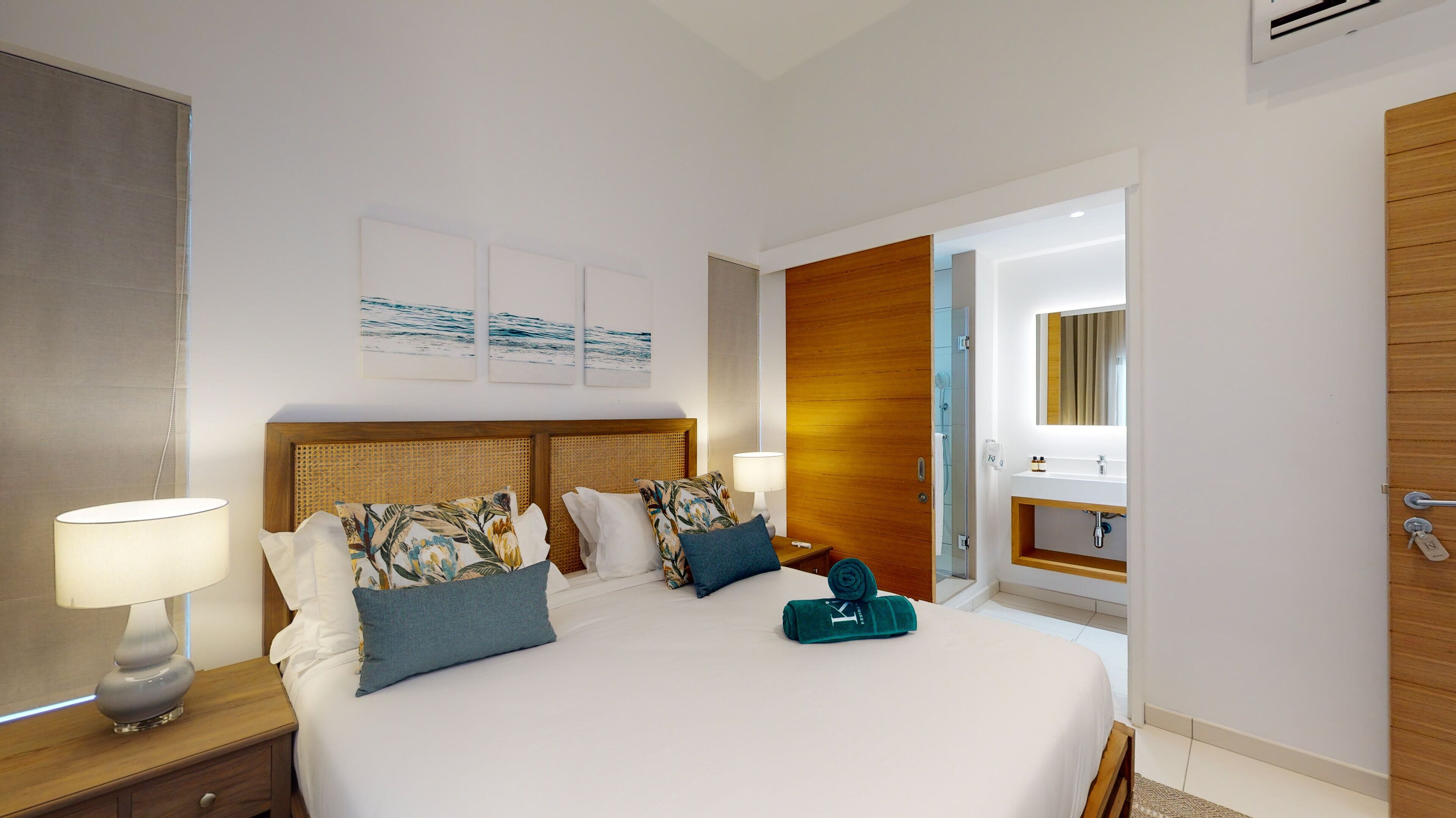 A hotel room with a large bed, two bedside tables with lamps, a wooden wardrobe, a bathroom with a mirror and sink, and artwork on the walls.