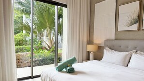 A hotel room with a bed, a nightstand with a lamp, a large window with curtains, and a view of greenery outside. -  Ki Residences with LOV (Grand-Baie)