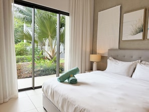 A hotel room with a bed, a nightstand with a lamp, a large window with curtains, and a view of greenery outside. -  Ki Residences with LOV (Grand-Baie)