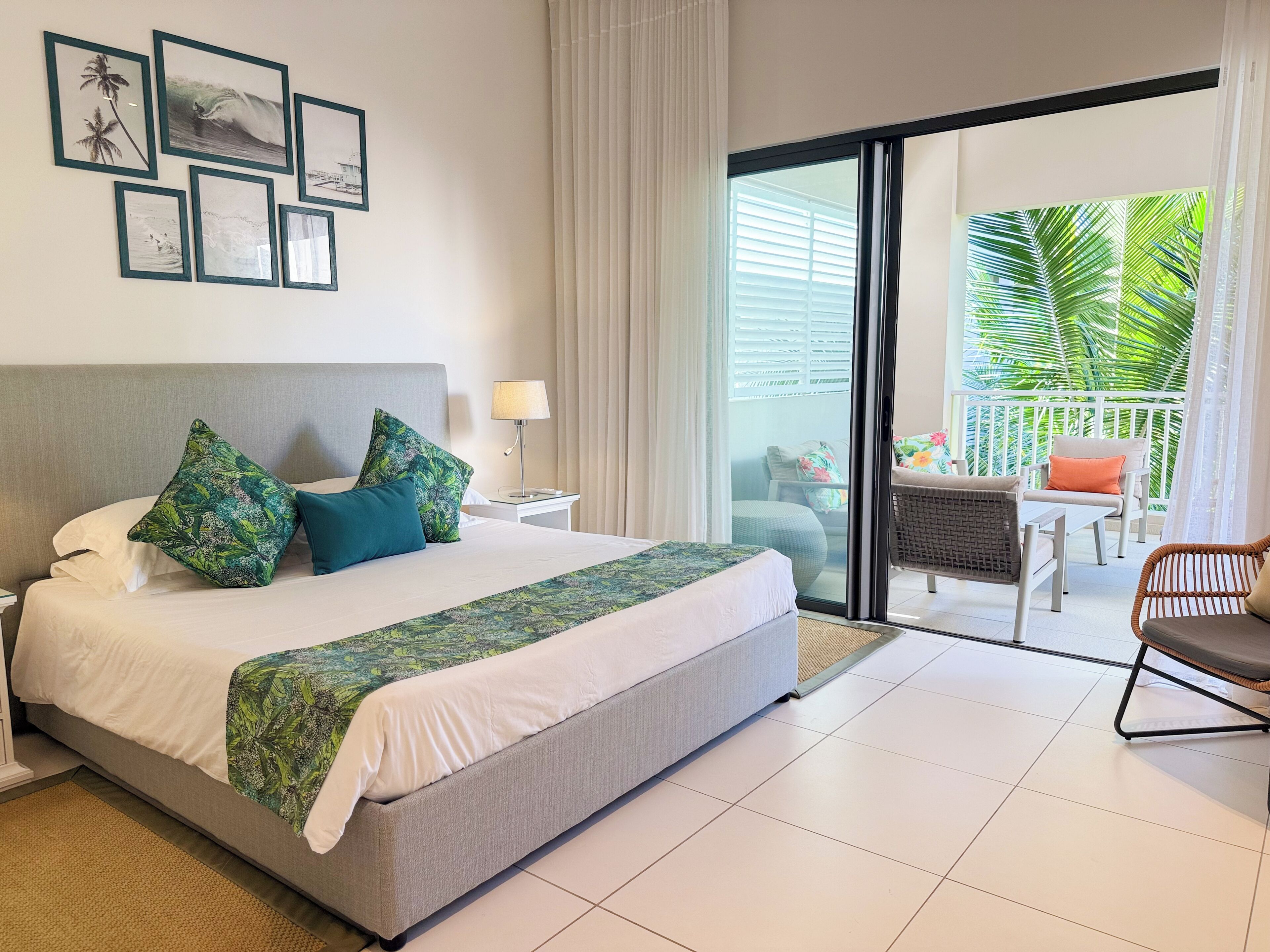 A hotel room with a bed, a nightstand, a lamp, a balcony with a chair, and a view of palm trees.