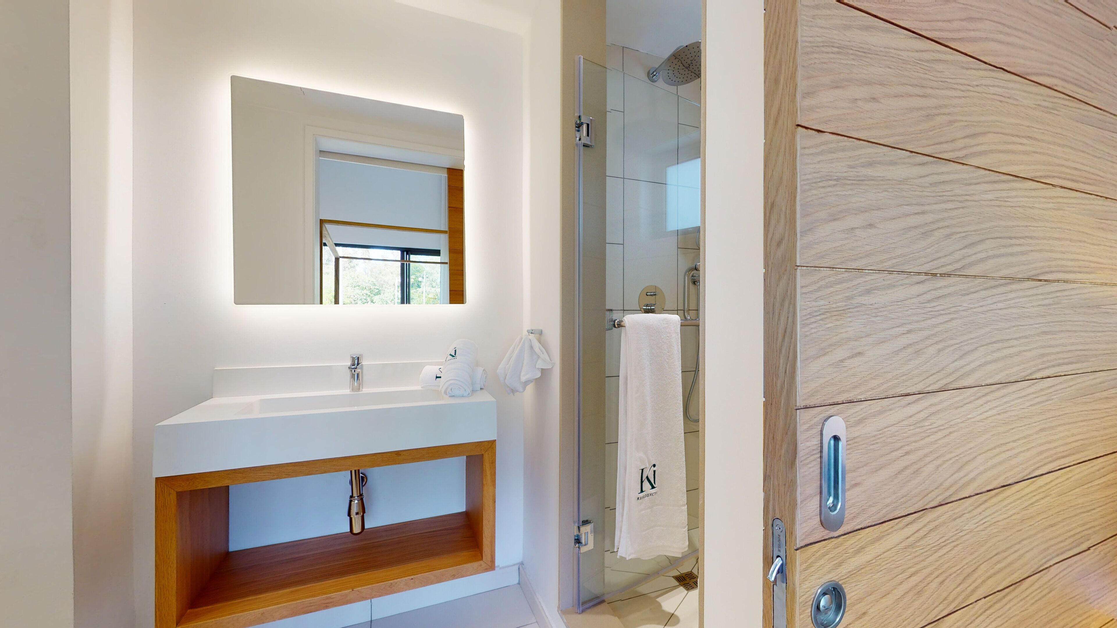 A modern bathroom with a white vanity, a glass shower enclosure, and a wooden cabinet with a towel hanging on the door.