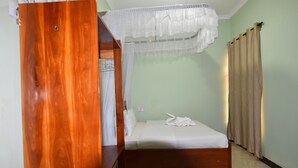 Een eenpersoonsbed met een wit laken en een handdoek, een houten garderobe, een witte hemel, beige gordijnen en een rood kleed.