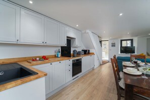 A modern kitchen with white cabinets, a wooden countertop, and a built-in oven. There is a dining area with a wooden table and chairs, and a TV mounted on the wall. - Burrow Cottage, Downalong – Sleeps 6 – Dog-Friendly (St Ives)