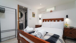 A neatly made bed with white linens and rolled towels, a wooden headboard, and a bedside table with a bottle and glasses.