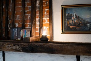 Un interior rústico con paredes de ladrillo visto, un estante de madera con libros, una vela encendida y un cuadro con una escena de pueblo.