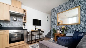 A modern kitchen with wooden cabinets, a built-in oven, and a black countertop. There is a dining area with a small table and chairs, and a large mirror on the wall. - Little Waves, St Ives – Sleeps 2 – Courtyard - Garage Parking (St Ives)