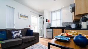 A modern living room with a grey sofa, a dining table with a blue vase, and a kitchen area with a refrigerator and a window. - Little Waves, St Ives – Sleeps 2 – Courtyard - Garage Parking (St Ives)