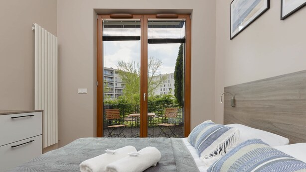 Habitación de hotel con cama, cabecera de madera, vista a un patio con mobiliario exterior y una puerta corrediza de vidrio que da al exterior.