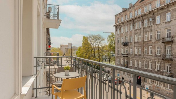 Un balcón con una mesa y sillas, con vista a una calle con edificios y un cielo despejado.