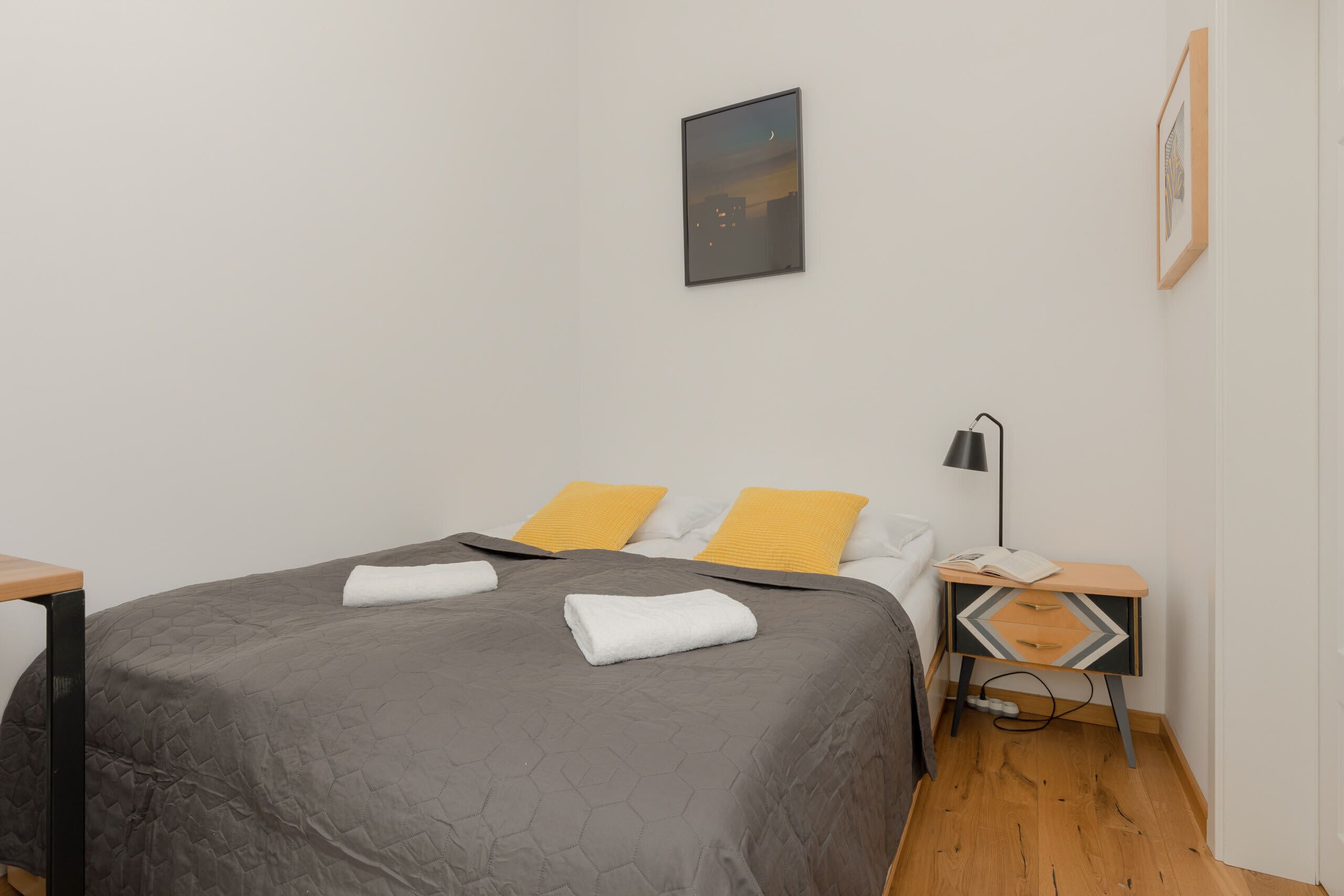 A neatly made bed with a gray quilt, two white pillows, and two yellow pillows. There is a framed picture on the wall above the bed and a small wooden nightstand with a lamp beside the bed.