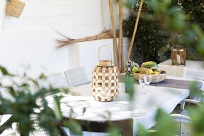 Una mesa redonda con mantel blanco, un farol tejido, una botella y una cesta de frutas.