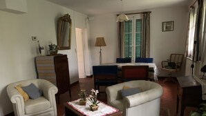 Ein Wohnzimmer mit einer Couch, Sesseln, einem Holzkabinett, einem Tisch mit Blumen, einer Lampe und einem Fenster mit Vorhängen.