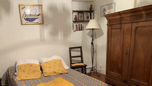 Ein ordentlich bezogenes Bett mit einem gemusterten Überwurf und zwei Kissen, ein Holzstuhl, eine Stehlampe und ein Holzschrank in einem Zimmer mit weißen Wänden und einem Fliesenboden.