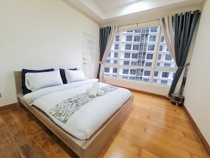 A neatly made bed with a floral bedspread and pillows, a wooden floor, a window with curtains, and a view of buildings outside.