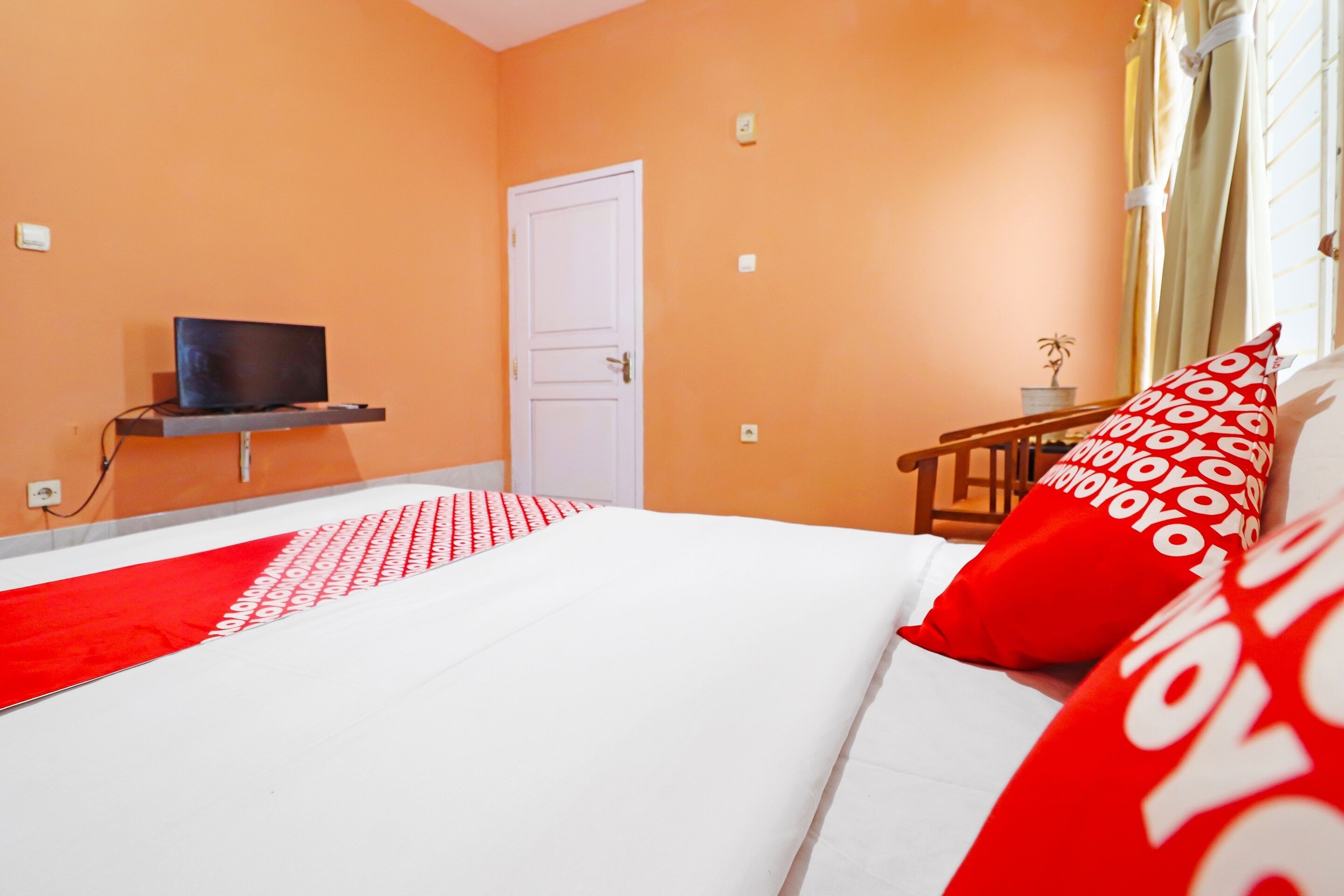 Un dormitorio con cama, televisor sobre un mueble, una puerta y una ventana con cortinas.
