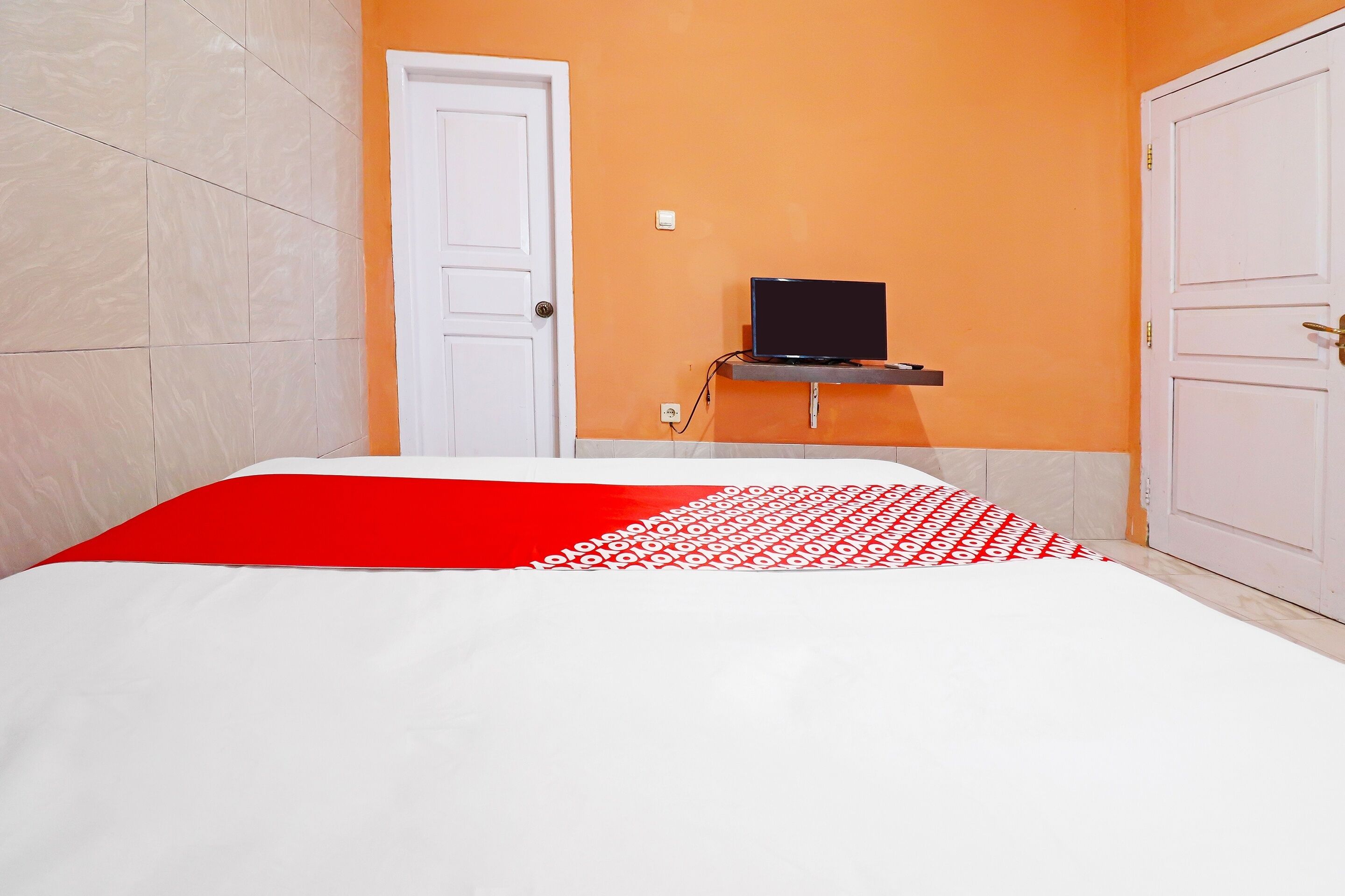 A bedroom with a bed, a white door, an orange wall, and a small shelf with a black object.