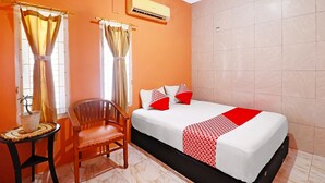 A hotel room with a bed, a chair, a small table with a plant, and an air conditioning unit.