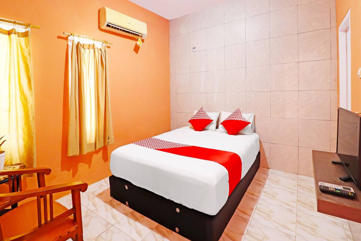 A hotel room with a bed, curtains, an air conditioning unit, a TV, and a remote control on a side table.