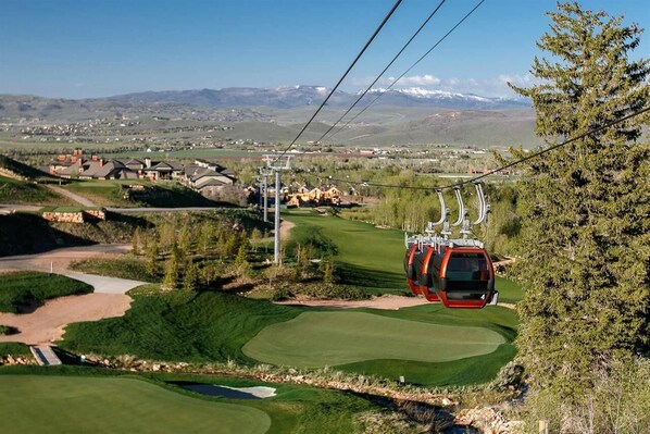 Un teleférico se desplaza sobre un campo de golf con montañas al fondo.