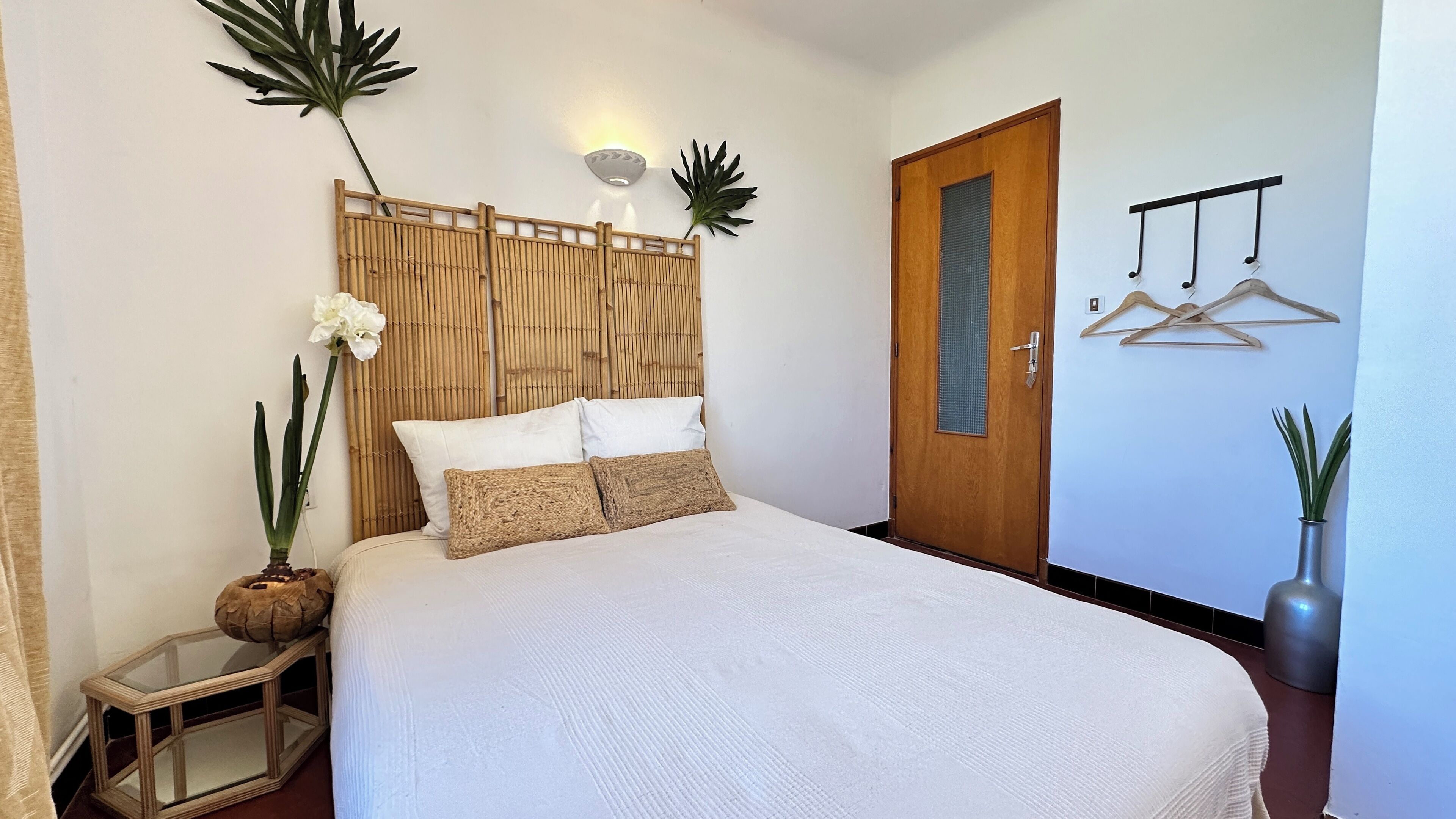 A neatly arranged bedroom with a bed, a wooden headboard, a bedside table with a mirror, and a door with a hanging coat rack.
