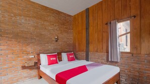 Una habitación con pared de ladrillo, paneles de madera, cama con ropa de cama blanca y detalles en rojo, ventana con cortinas y un estante con una cesta.