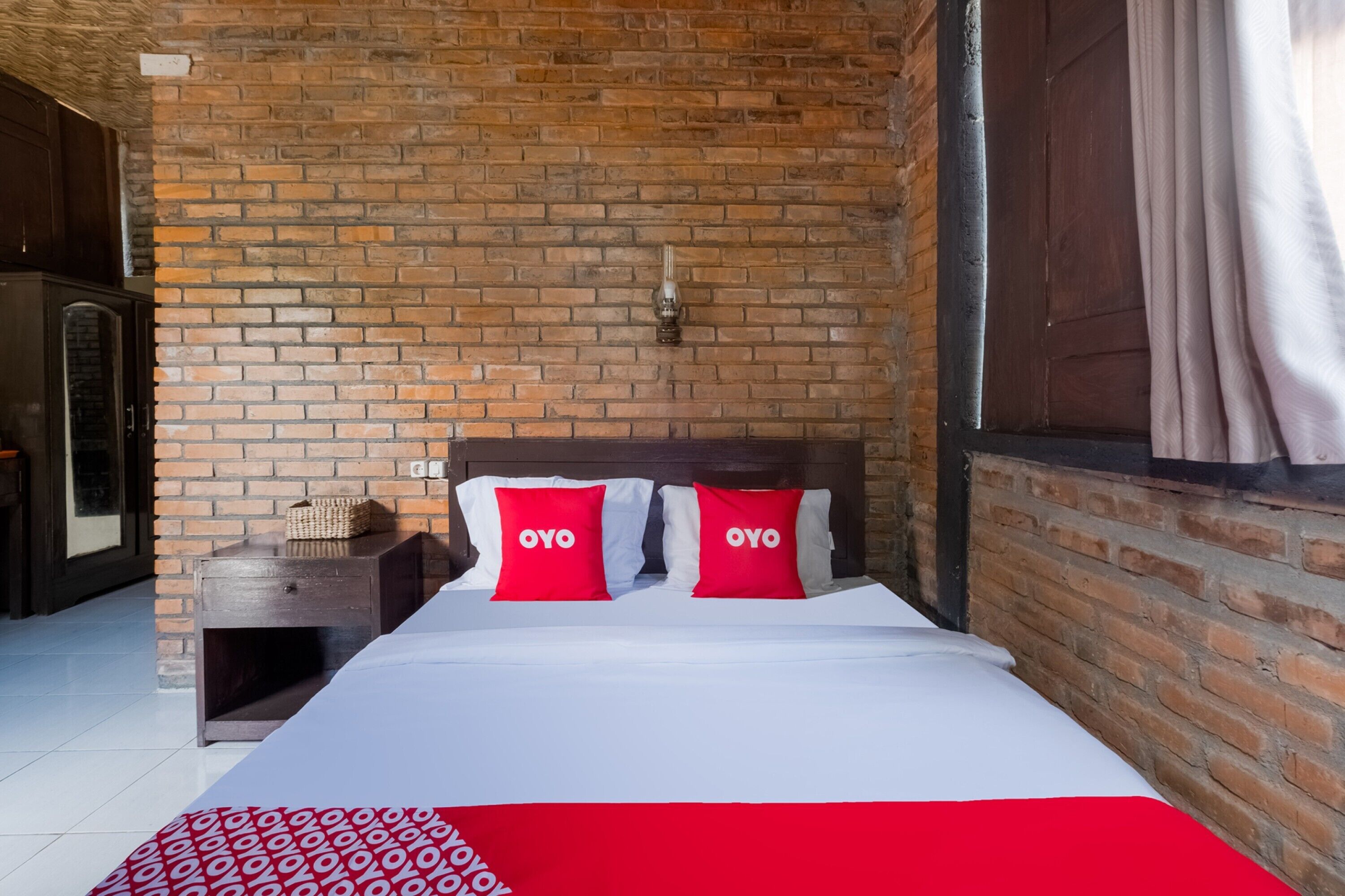 A hotel room with a brick wall, a wooden headboard, a bed with white bedding and red pillows, and a nightstand with a basket.