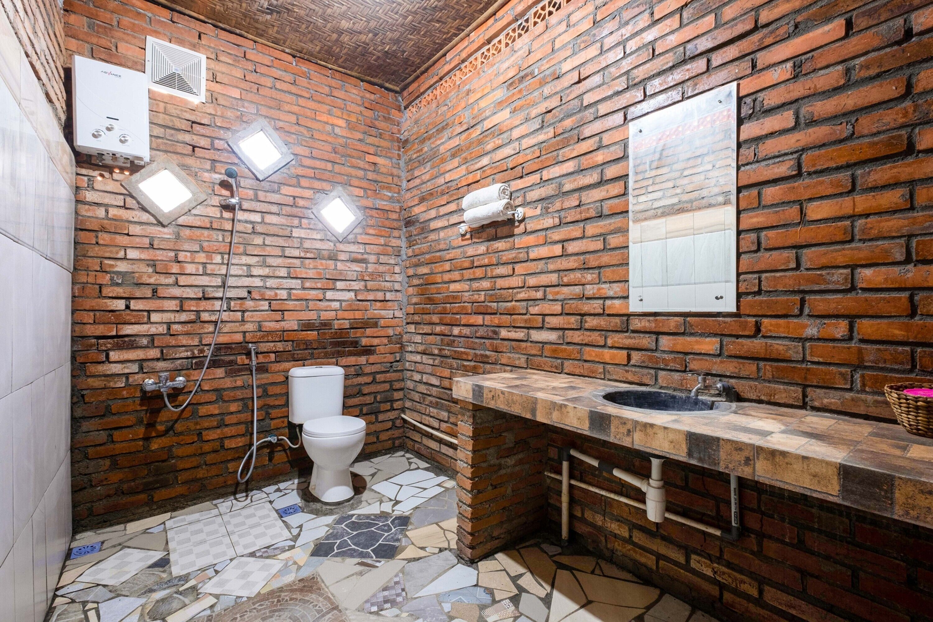 Un baño con pared de ladrillo, inodoro blanco, lavabo con encimera de mosaico y zona de ducha.