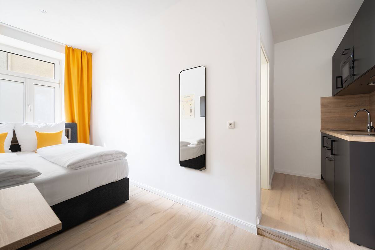 Une chambre d’hôtel moderne équipée d’un lit, d’une table de chevet, d’un miroir, d’une fenêtre avec des rideaux jaunes et d’une kitchenette comprenant un évier et un four à micro-ondes.