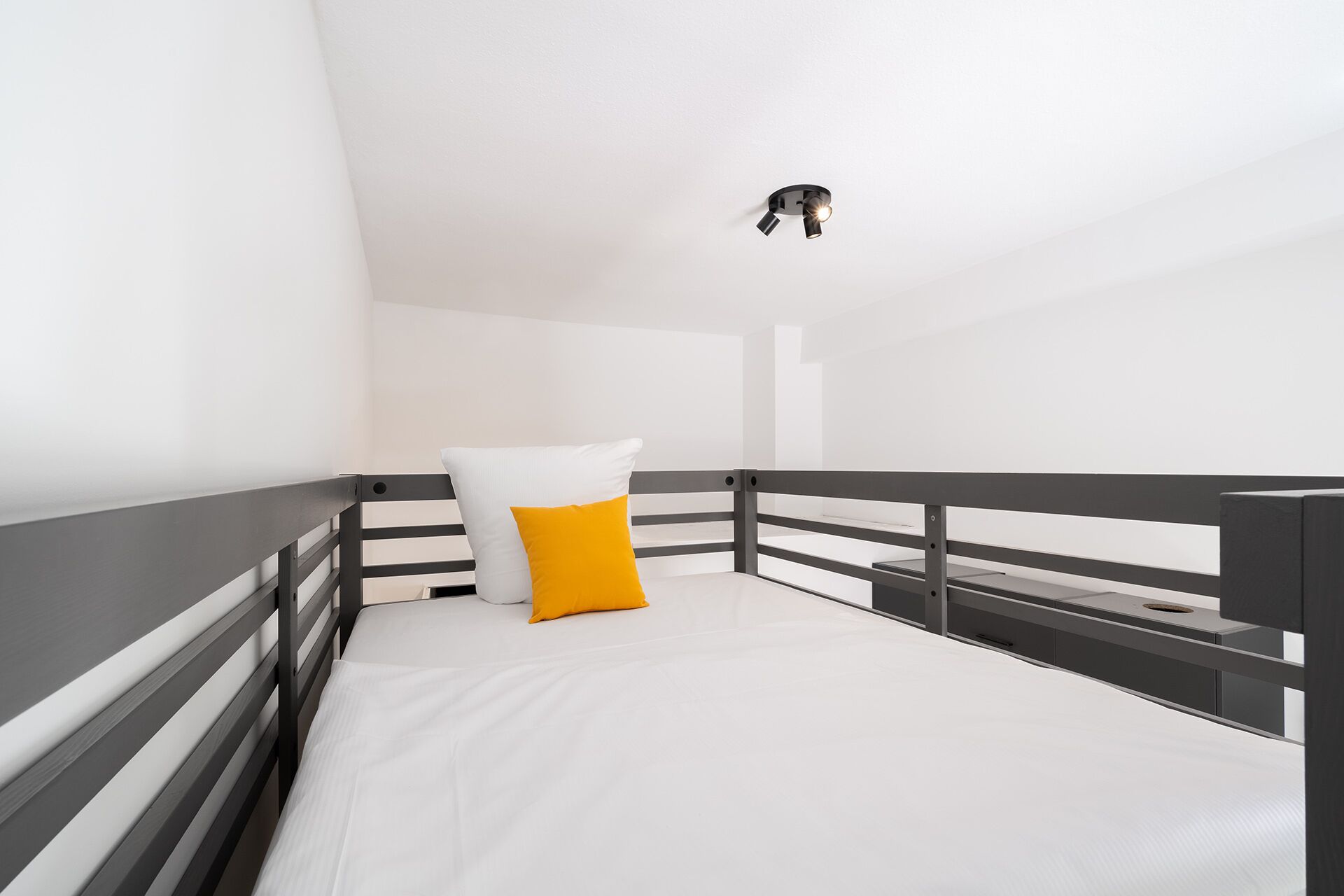 Una cama individual con colchón blanco y almohada amarilla, barandal negro y una lámpara en el techo.
