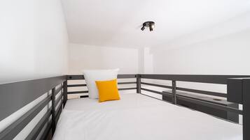Una cama individual con colchón blanco y almohada amarilla, barandal negro y una lámpara en el techo.