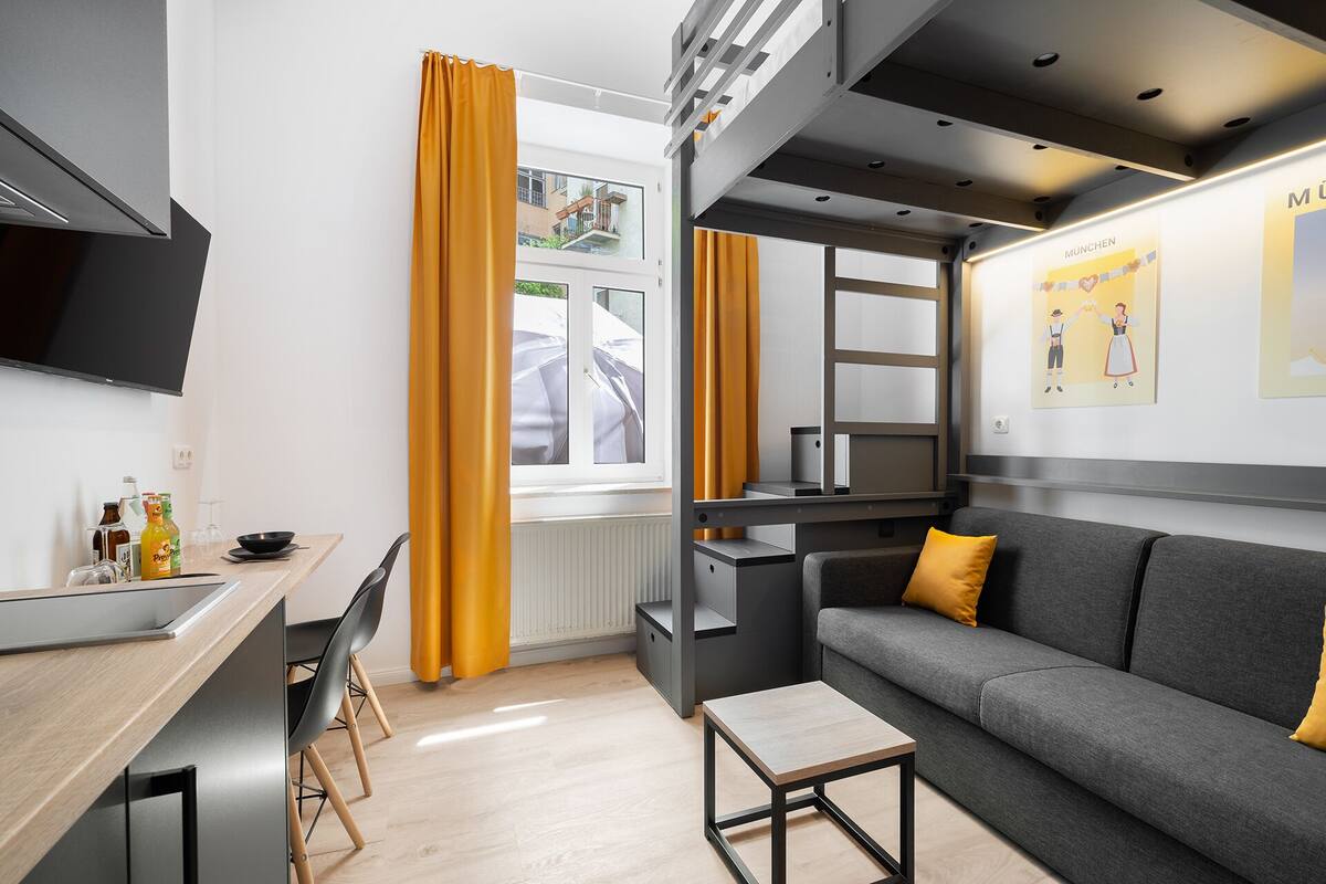 Une chambre d’hôtel moderne avec un canapé gris, une petite table en bois et un coin cuisine avec une fenêtre.