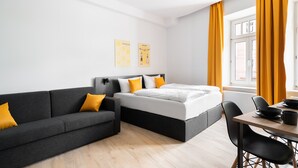 A modern hotel room with a grey sofa, a bed with white bedding, and a dining area with a table and chairs. - Servus Apartments by Homaris (Munich)
