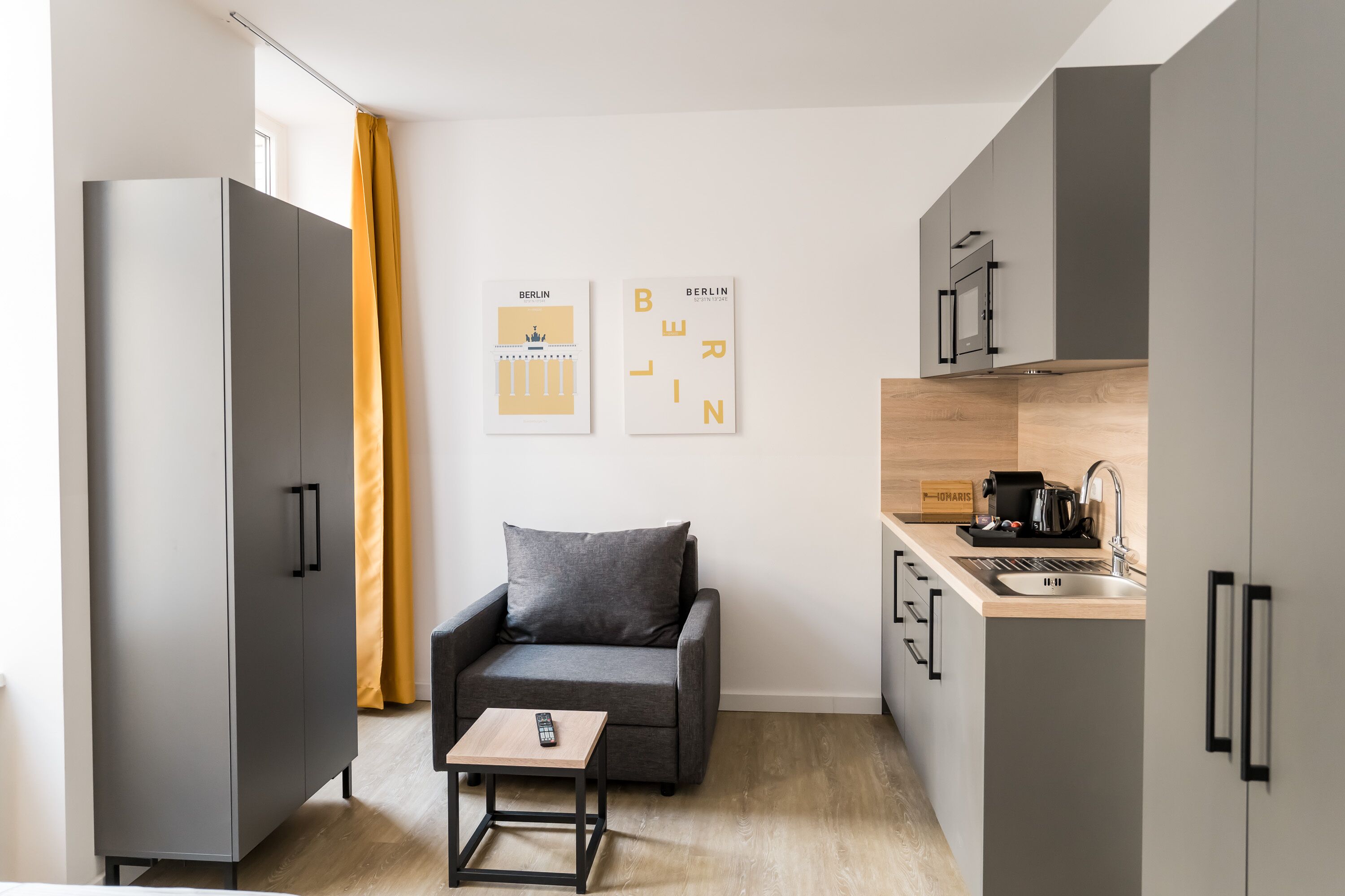 Un espace de vie moderne et compact, avec un canapé gris, une petite table, un coin cuisine équipé d’un évier et d’une machine à café, et deux tableaux encadrés accrochés au mur.