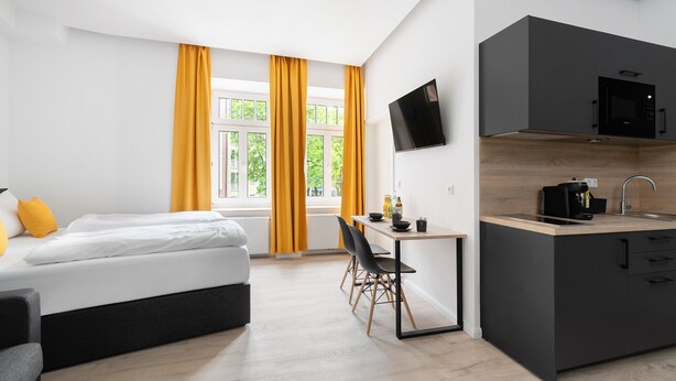 A modern hotel room with a bed, a desk, a chair, a TV, and a kitchen area. - Servus Apartments by Homaris (Munich)