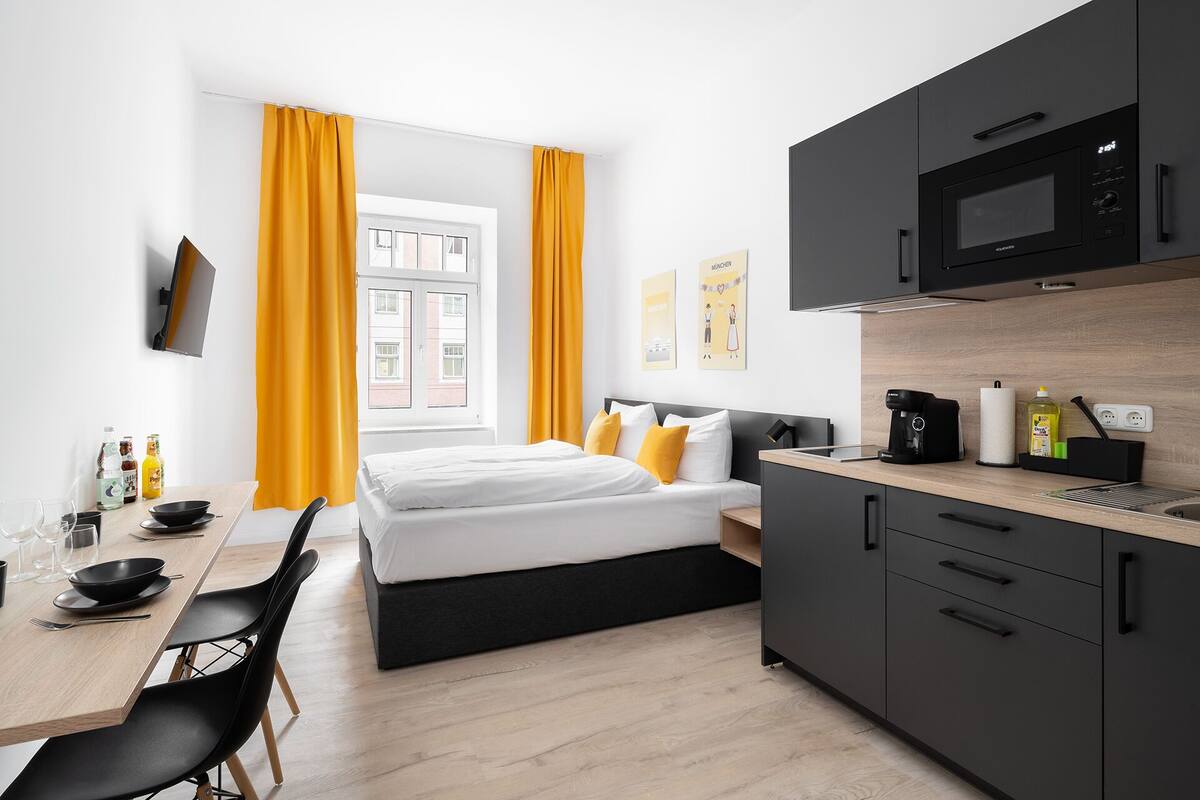 Une chambre d’hôtel moderne comprenant un lit, une table à manger avec des chaises, un coin cuisine avec un four à micro-ondes et une fenêtre avec des rideaux.