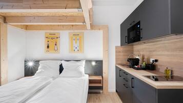Ein Schlafzimmer mit einem Bett, einem Nachttisch und einer Holzbalkendecke. Es gibt eine Küchenzeile mit einer Mikrowelle, einer Kaffeemaschine und einem Spülbecken.