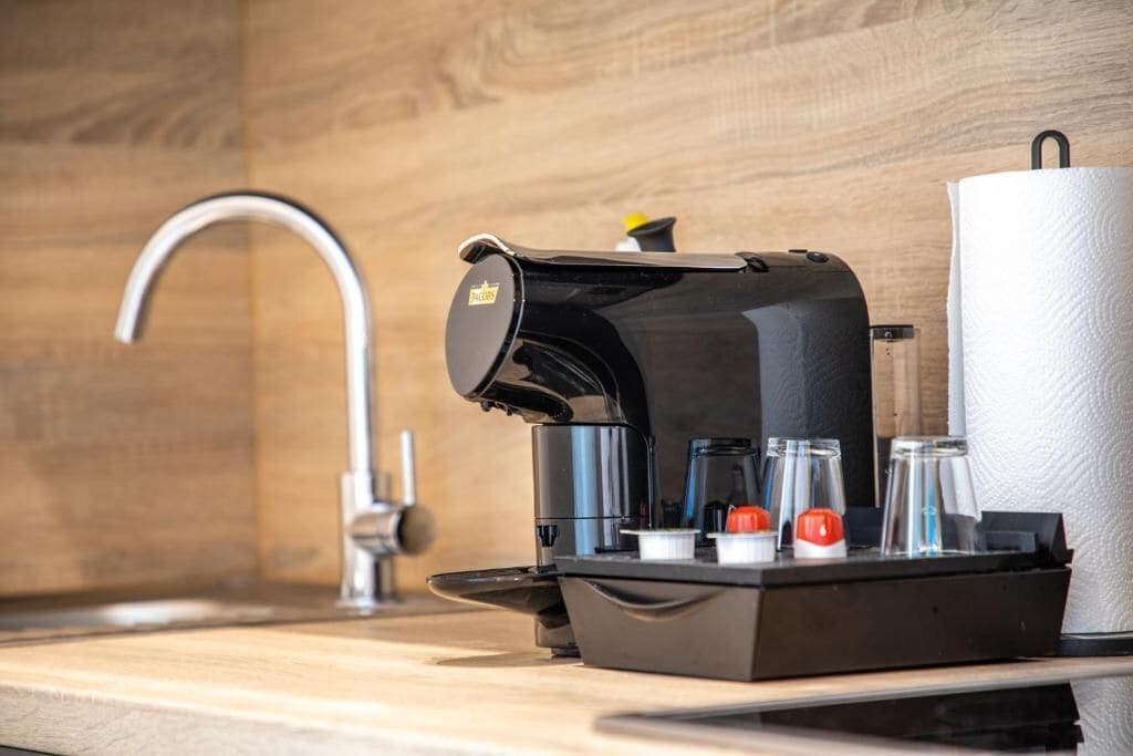 Una cafetera moderna con depósito de agua, cápsulas de café y dispensador de toallas de papel sobre una encimera de madera.