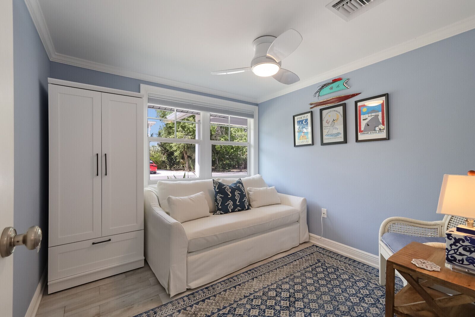 A room with a white sofa, a ceiling fan, a window with a view of the outside, a patterned rug, and a small table with a lamp.