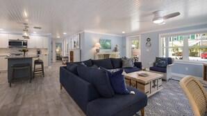 Un vaste espace de vie avec un canapé bleu foncé, une table basse en bois et un fauteuil en osier. On aperçoit au fond une cuisine avec des meubles blancs et un four à micro-ondes.
