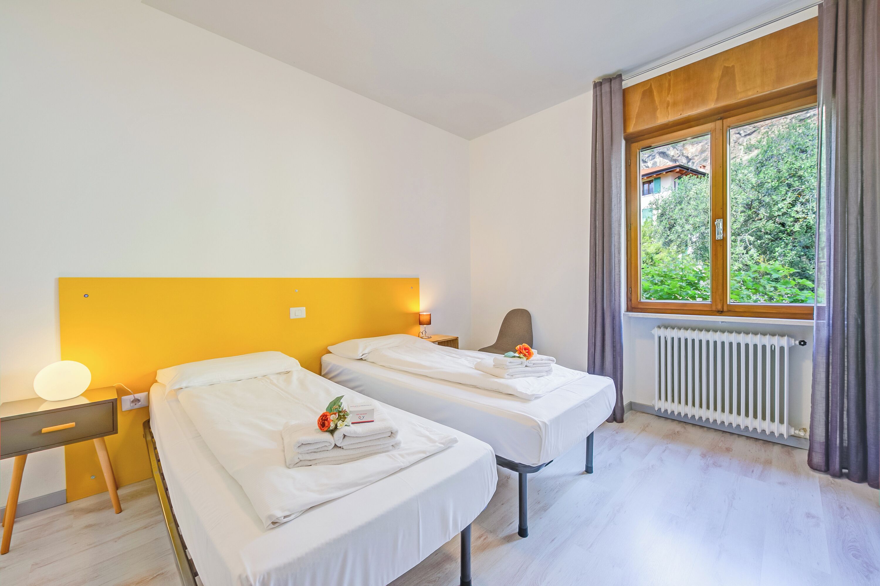 Een hotelkamer met twee bedden, een nachtkastje, een lamp, een raam met gordijnen en een radiator.