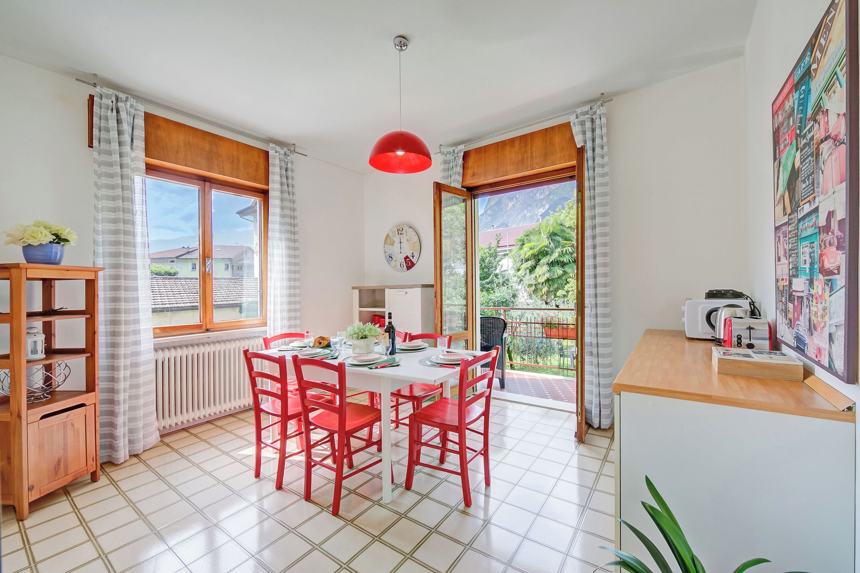 A dining area with a table, chairs, a red pendant light, a clock, and a view of greenery outside.