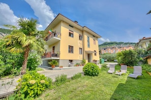 A two-story yellow building with a balcony, surrounded by greenery and a paved courtyard.