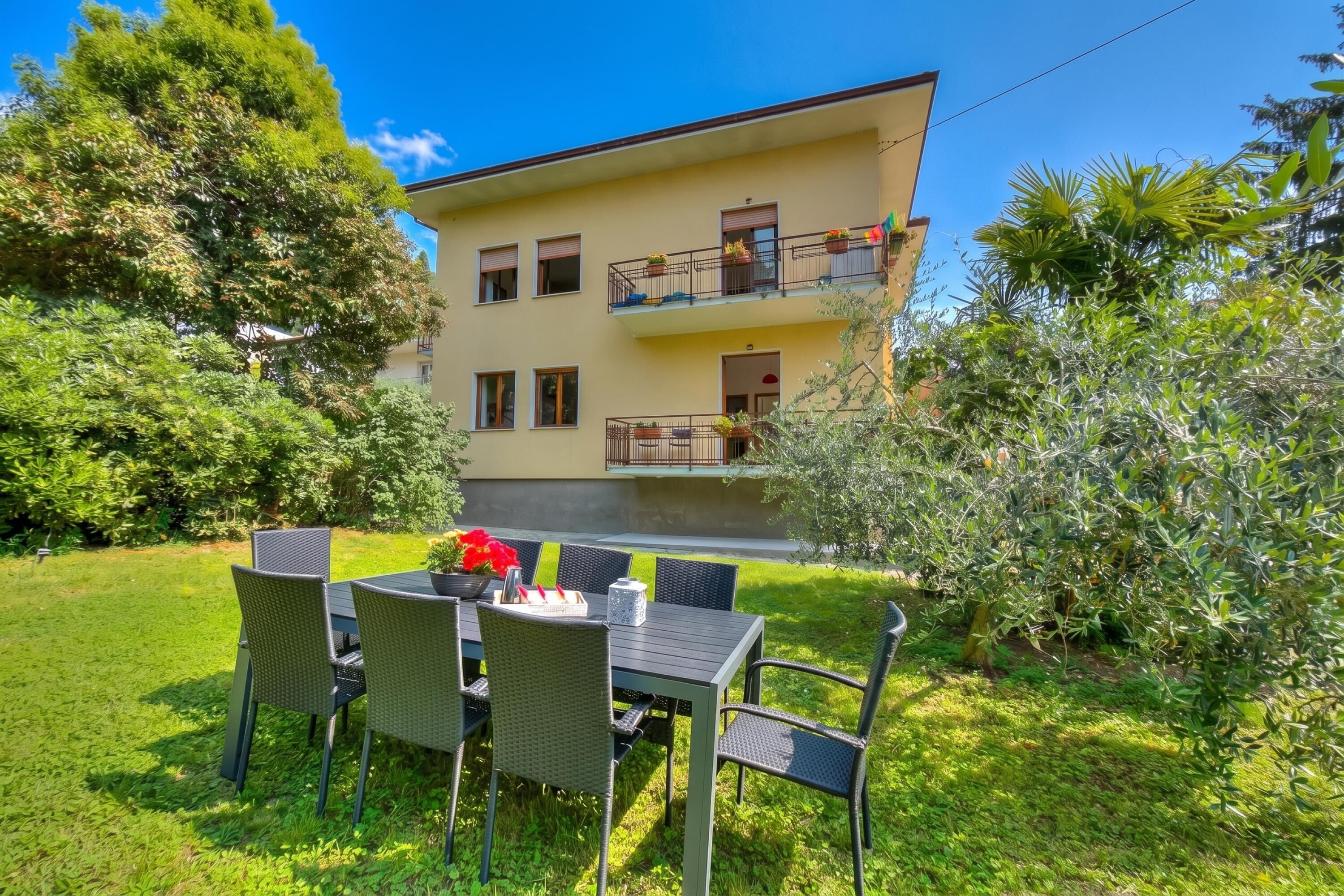 A two-story building with a balcony, a garden with a table and chairs, and a clear blue sky.