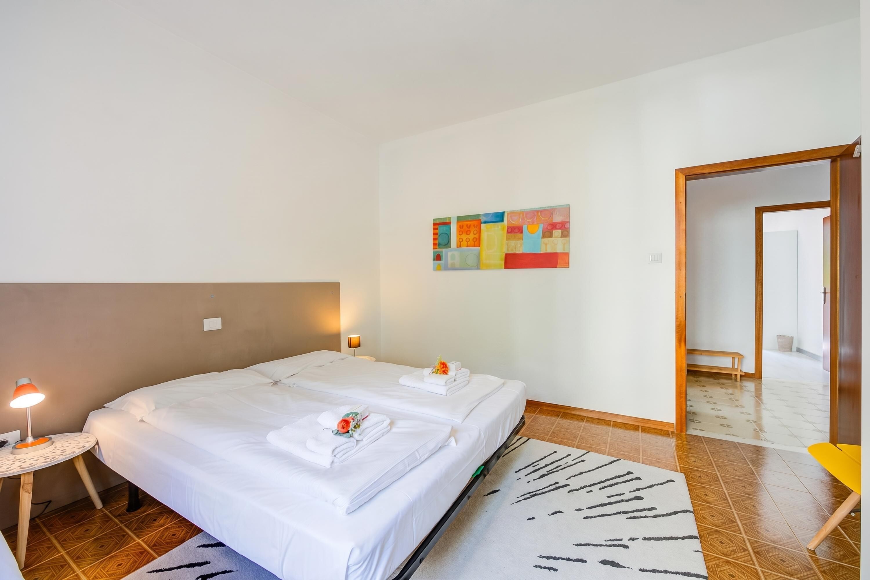 A hotel room with two beds, a small table with a lamp, a colorful abstract painting, and a wooden door.