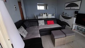 Un salon moderne avec un canapé d’angle, un téléviseur à écran plat et un coin repas comprenant des chaises blanches et une table.