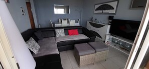 A modern living room with a sectional sofa, a flat-screen TV, and a dining area with white chairs and a table.