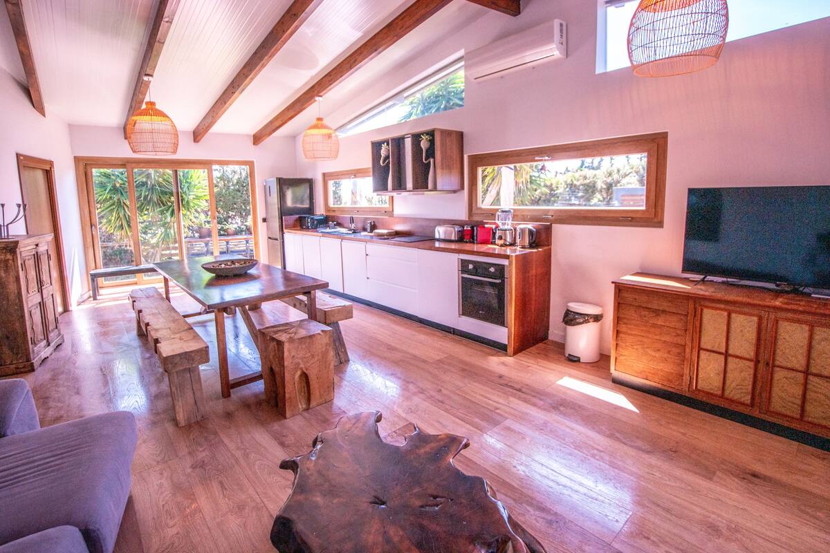 A spacious kitchen with wooden countertops, a dining area with benches, and a large TV on the right.