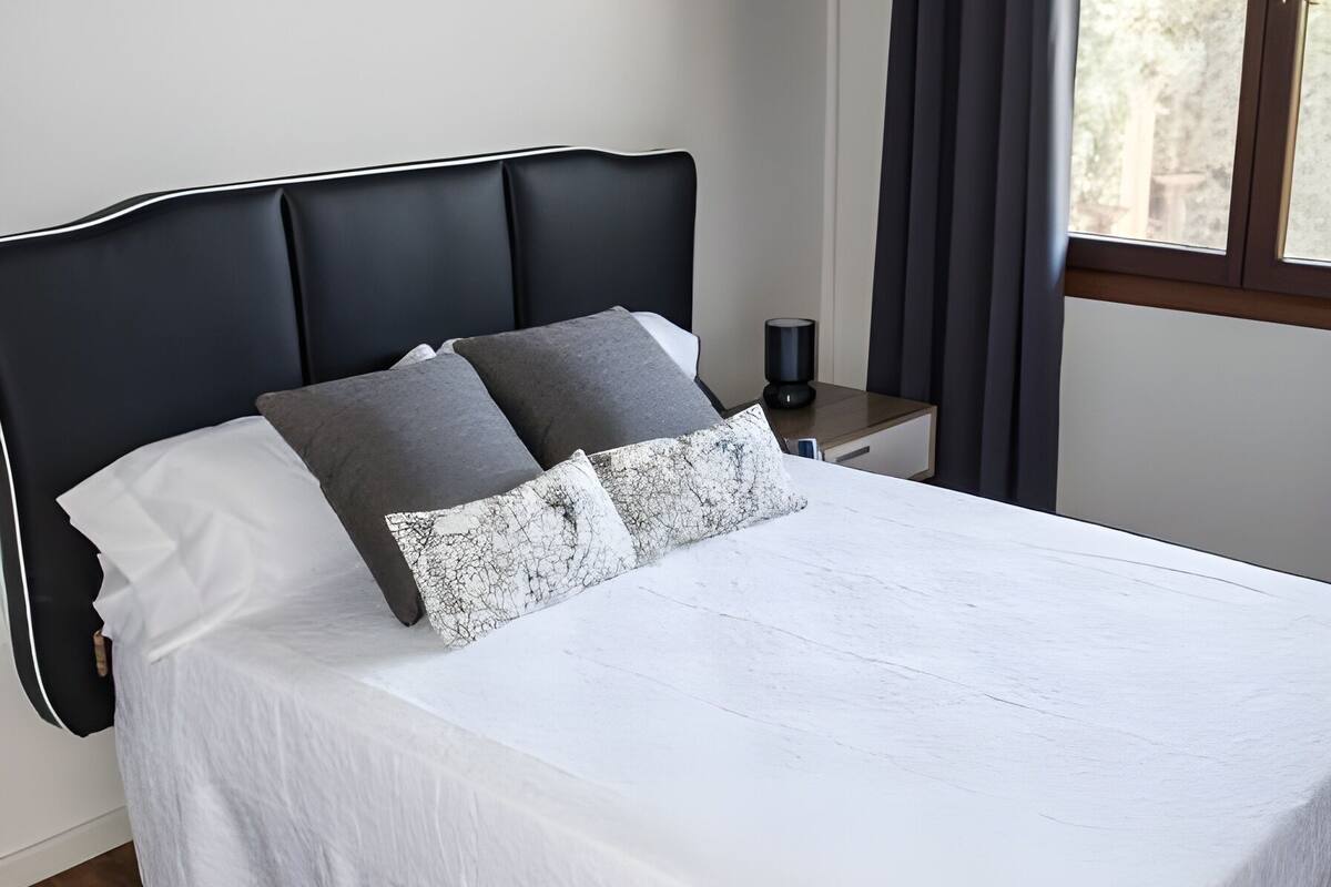 A neatly made bed with a white bedspread and multiple pillows, a wooden floor, a dark headboard, and a window with curtains.