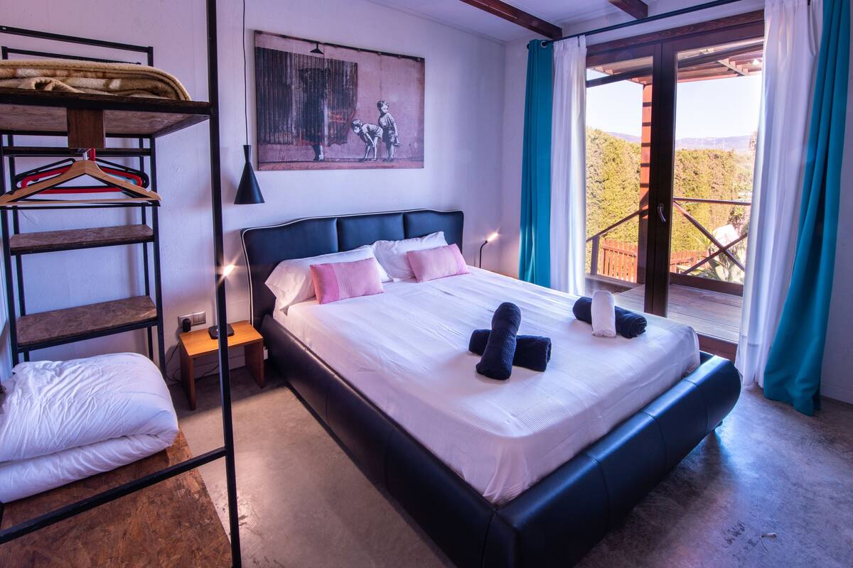 A bedroom with a bed, a wooden shelf with clothes, a painting on the wall, and a balcony with a view.