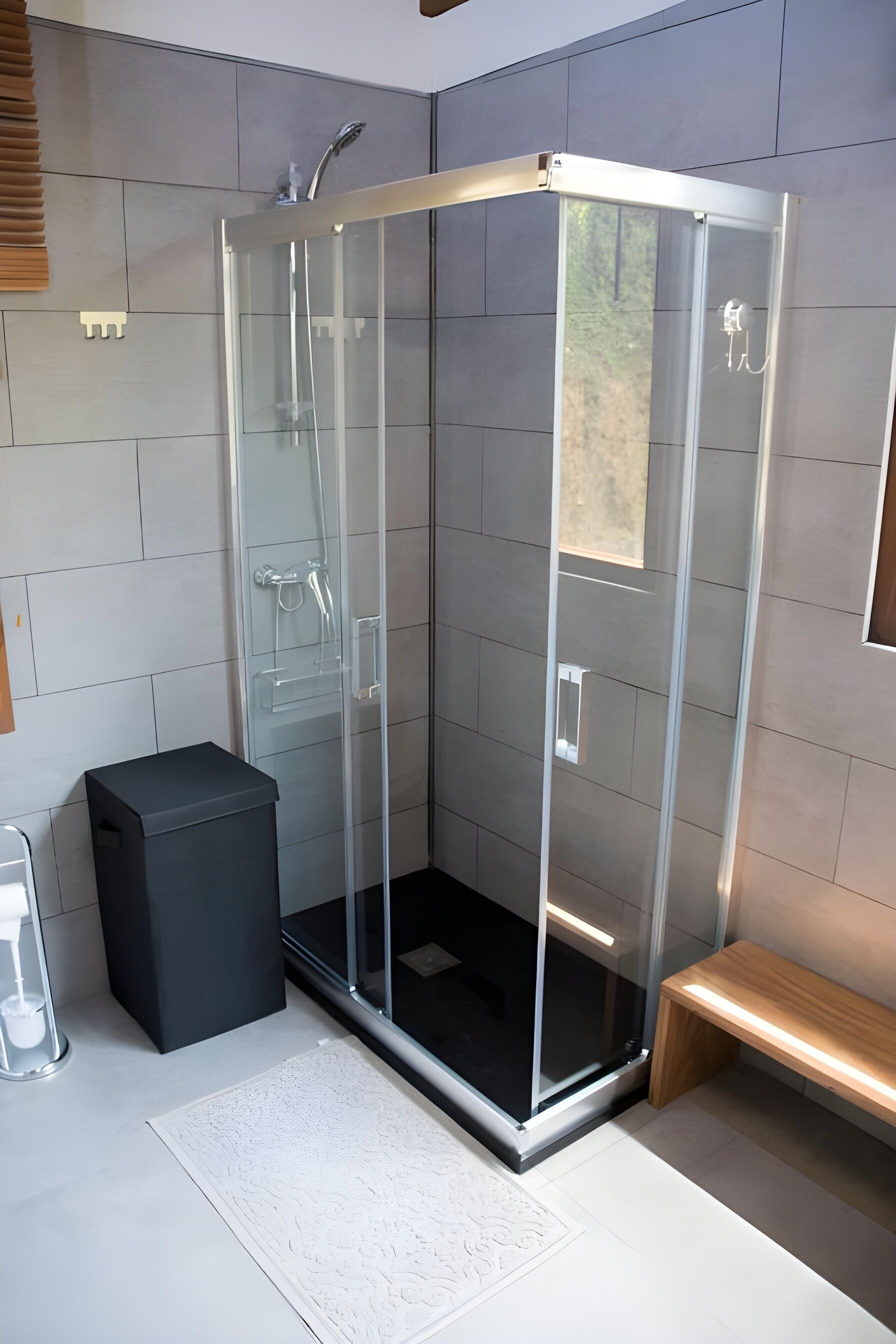 Un baño moderno con ducha envuelta en vidrio, un cubo de basura negro, un banco de madera y un tapete blanco con textura.